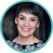 Une femme aux cheveux sombres et courts, vêtue d’un chemisier à collier de fleurs, sourit.