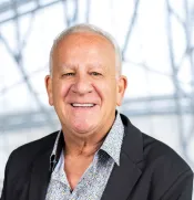 Un homme blanc aux cheveux blancs courts porte un blazer noir, superposé à une chemise à motifs. Il est de face et sourit à l’appareil photo.