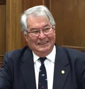 Bill Shead est un homme d’un certain âge, aux cheveux blancs et aux lunettes. Il sourit, regarde vers le côté et porte un costume bleu foncé et une cravate bleue ornée d’avions de chasse blancs.