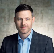 Un homme blanc vêtu d'une veste de costume à carreaux et d'une chemise bleue pose pour une photo professionnelle sur un fond de pierre. Il a des cheveux bruns courts et une barbe taillée.