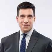 Un homme blanc aux cheveux foncés sourit légèrement à la caméra. Il porte un costume sombre avec une chemise blanc cassé et une cravate bleu foncé à petits pois blancs.