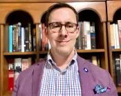 Un homme blanc aux cheveux bruns et portant des lunettes sourit à la caméra. Il porte un blazer violet avec une épingle à la boutonnière et une chemise multicolore.