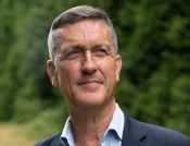 Un homme blanc aux cheveux gris courts regarde vers le haut, hors champ. Il porte un blazer bleu et une chemise à rayures bleues.