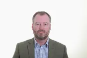 Un homme blanc aux cheveux grisonnants courts et à la barbe regarde la caméra d’un air impartial. Il porte un blazer gris et une chemise à carreaux bleus et blancs.