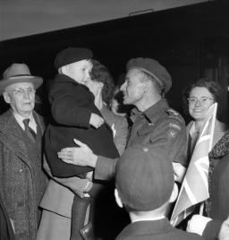 Photo noir et blanc d’un homme portant un uniforme de l’armée canadienne, qui sourit et parle à un petit enfant dans les bras d’une femme. D’autres hommes et d’autres femmes sont rassemblés autour.