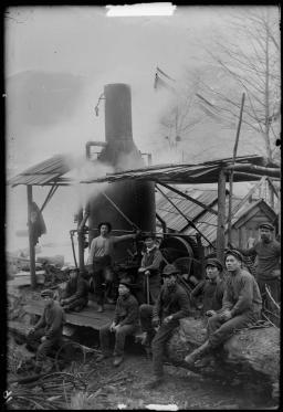 Une photographie en noir et blanc de neuf hommes devant un camp de bûcherons. Trois hommes sont debout et les autres sont assis.