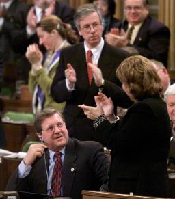 Dans la Chambre des communes, un homme est assis alors que ses collègues politiciens sont debout et applaudissent.
