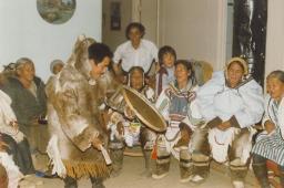 A man dancing and playing Un homme danse en jouant un tambour inuit alors qu’un groupe de femmes plus âgées chante. Inuit drum while a group of elderly women sings.