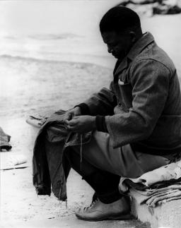 Une image en noir et blanc de Nelson Mandela. Il est assis et tient un vêtement sur ses genoux. Il semble être en train de coudre. Il porte des shorts, de longues chaussettes, des chaussures blanches et un veston.