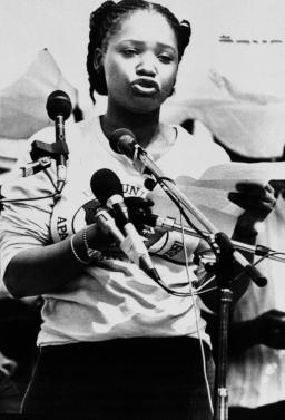 Une image en noir et blanc de Zindzi Mandela, une jeune femme noire, debout et prenant la parole devant plusieurs microphones.