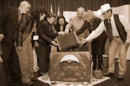 Un groupe de personnes entoure la boîte de bois cintré de la Commission de vérité et réconciliation. Les personnes sont penchées vers la boîte et tendent la main pour placer une valise dans la boîte.