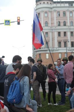 Deux soldats en uniforme sont debout derrière une foule de jeunes gens. Deux des jeunes tiennent un drapeau aux rayures rouge, blanche et bleue. On voit un haut bâtiment en arrière-plan.