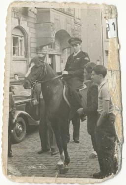 Photo en noir et blanc aux bords abimés d’un homme à cheval en uniforme dans la rue. Deux hommes se tiennent à côté. On voit une vieille voiture à l’arrière-plan.