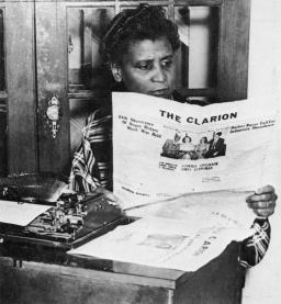 Une image en noir et blanc d’une femme noire assise devant une vieille machine à écrire, lisant un journal intitulé « The Clarion ».
