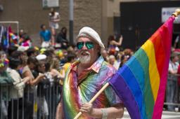 Un homme portant des lunettes de soleil et une chemise aux couleurs de l’arc-en-ciel porte un grand drapeau arc-en-ciel tandis qu’une foule de personnes le regardent de loin.