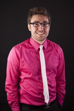 Un jeune homme portant une chemise rose, une cravate blanche et des lunettes noires sourit à l’objectif.