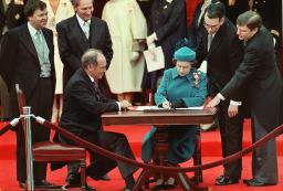 L’ancien premier ministre Pierre Elliott Trudeau est assis à une table en bois à la gauche de la reine Elizabeth II. Deux hommes se penchent et regardent au-dessus de l’épaule de la reine alors qu’elle signe un document.