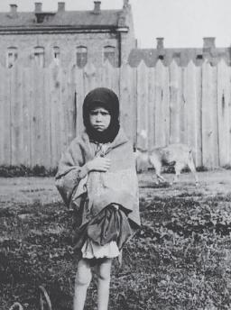 Une jeune fille se tient face à la caméra avec une expression de tristesse, serrant un châle autour d’elle.