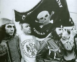 Quatre hommes portant des uniformes de fortune se tiennent debout avec un drapeau noir orné d’une tête de mort.