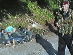 Image fixe granuleuse tirée d’une vidéo montrant un homme portant un uniforme de camouflage au premier plan. Plusieurs personnes en civil sont allongées face contre terre, les mains liées.