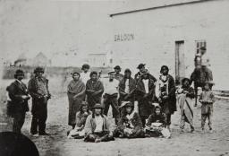 Photographie en noir et blanc d’un groupe d’hommes debout et assis devant un bâtiment avec le mot « Saloon » peint en évidence sur le mur.