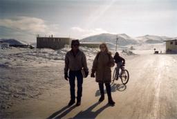 Deux personnes portant un manteau d’hiver se tiennent dans un paysage enneigé, devant des collines et des bâtiments en arrière-plan.