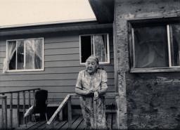 Une femme aux cheveux blancs s’appuie sur le garde-fou du balcon à l’extérieur d’une maison. 