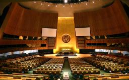 Une grande salle avec des rangées de bureaux et de chaises faisant face à un haut mur orné du logo de l’ONU.