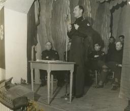 Un homme portant un uniforme orné d’une croix gammée parle au microphone sous les yeux d’un groupe d’hommes portant un uniforme semblable.