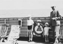 Un homme vêtu d’un complet pose avec deux enfants contre la rambarde d’un navire. 