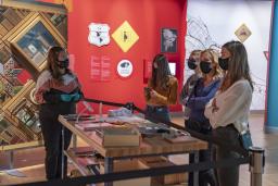 Une femme portant des gants bleus est debout derrière une table couverte d’objets et parle à quatre jeunes femmes de l’autre côté de la table.