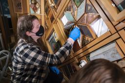 Une personne aux cheveux longs, portant un masque et des gants bleus, nettoie une section d’une œuvre d’art à cadre en bois avec une petite éponge.