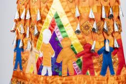 Une section d'une robe orange à clochettes est représentée. Les clochettes, ou petits pendentifs argentés en forme de cône, sont visibles en haut de l'image. Sous elles, sept figures humaines sont représentées, toutes de couleurs et de tailles différentes. L'une des figures porte une chemise orange. Ces figures humaines se tiennent la main à l'intérieur d'un tipi multicolore.