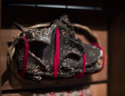 Une vieille chaussure d’enfant en cuir, abîmée et couverte de lichens. Elle a été enveloppée d’une bande de tissu rouge, montée sur un support en bois et entourée d’une tresse de foin d’odeur.