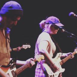 Deux guitaristes, l’un portant une tuque et l’autre une casquette de baseball, jouent sur scène sous une lumière violette. Un microphone et une autre guitare sont visibles en bordure.