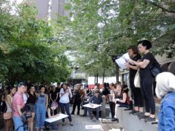 Une foule de quelques dizaines de personnes rassemblées dans un petit parc urbain écoutant deux personnes parler. Les personnes qui parlent sont debout sur un muret et tiennent des documents. Certains membres de la foule dessinent des pancartes de protestation.