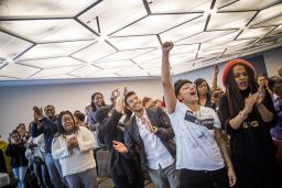 Une salle de conférence remplie de personnes noires de genres et d’âges différents, applaudissant et souriant avec enthousiasme, certaines levant le poing en l’air et faisant des signes de la main.