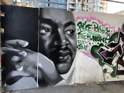 Grande fresque murale composée d’un portrait blanc, gris et noir du visage de Martin Luther King à côté d’un texte vert stylisé dans un contour rose déchiqueté, qui se lit « Police Brutality ENDS NOW! RIP Jamal » (Fin aux brutalités policières MAINTENANT! RIP Jamal).
