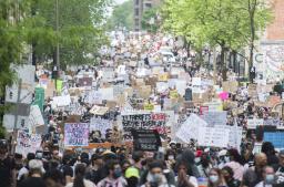 Une foule nombreuse et diversifiée remplit une large rue bordée d’arbres. Beaucoup brandissent des pancartes de protestation portant des slogans en anglais et en français tels que « Pas de justice, pas de paix », « Les vies noires comptent » et « Suis-je prochain·e? ».