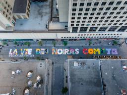 Photo aérienne d’une rue de centre-ville entre des immeubles de bureaux. La rue a été peinte avec un texte très grand et coloré indiquant « LA VIE DES NOIR.E.S COMPTE » avec un texte blanc plus petit indiquant « #BLACKLIVESMATTER ».