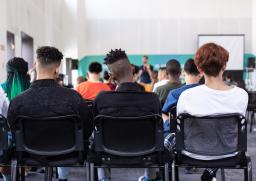 Un groupe de jeunes gens dans une salle de classe, vus de dos. Ils ont des couleurs de peau, des coiffures et des vêtements variés. 