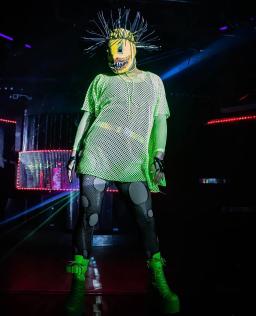 Une drag queen portant un masque jaune aux dents acérées, un haut et des bottes en maille vert vif, et des collants gris foncé avec de grands cercles gris clair.