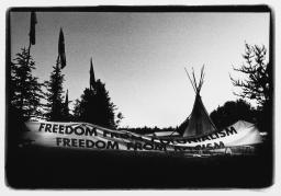 Photo en noir et blanc montrant une grande bannière portant l’inscription « Freedom from colonialism / Freedom from racism » (Liberté face au colonialisme / Liberté face au racisme) installée devant trois drapeaux et un tipi, dans un paysage forestier.