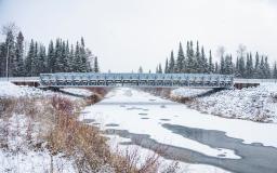 Un pont en métal enjambe un canal ressemblant à une rivière. L’eau est en grande partie gelée, il y a de la neige sur le sol et, à l’arrière-plan, on aperçoit de grands conifères. 
