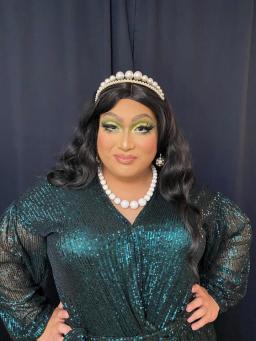 Une drag queen portant une robe vert foncé chatoyante, un diadème de perles et un collier de perles blanches.