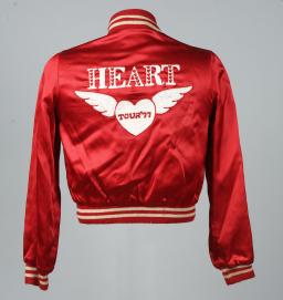 Le dos d’une veste en satin rouge vif avec le texte blanc « HEART » en grandes lettres majuscules au-dessus d’un cœur blanc ailé sur lequel est écrit « TOUR '77 ».