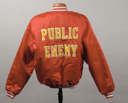 Le dos d’une blouson style aviateur de couleur rouge-orange foncé, ornée de l’inscription « Public Enemy » en grandes lettres majuscules jeunes.