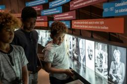 Trois enfants regardent une exposition dans un musée. L’exposition montre une série de photos en noir et blanc présentées en une longue rangée le long d’un mur. Des panneaux de texte de différentes tailles et couleurs sont suspendus au-dessus des photos.