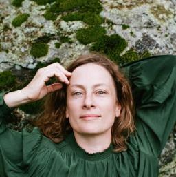 Une femme aux cheveux roussâtres et aux yeux verts, vêtue d’une chemise vert émeraude, est allongée sur un rocher recouvert de lichen. Une main est derrière sa tête, l’autre touche son front.