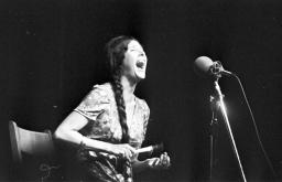 Une femme aux longs cheveux tressés, assise sur une chaise, chante dans un microphone tout en jouant d’un petit hochet.